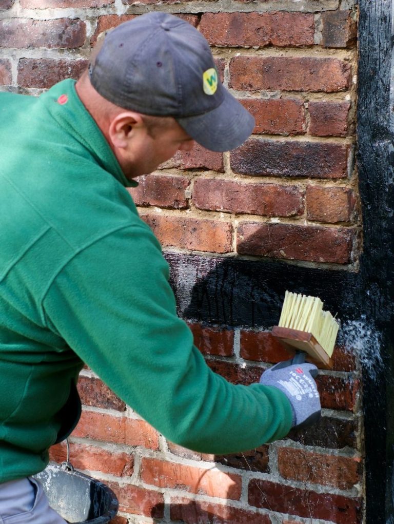 Kontrolliertes Vornässen der Klinkerwand vor dem Verfugen Handwerker im grünen Pullover befeuchtet mit Quast und Wasser die vorbereiteten Mauerwerksfugen an einer Backsteinfassade.