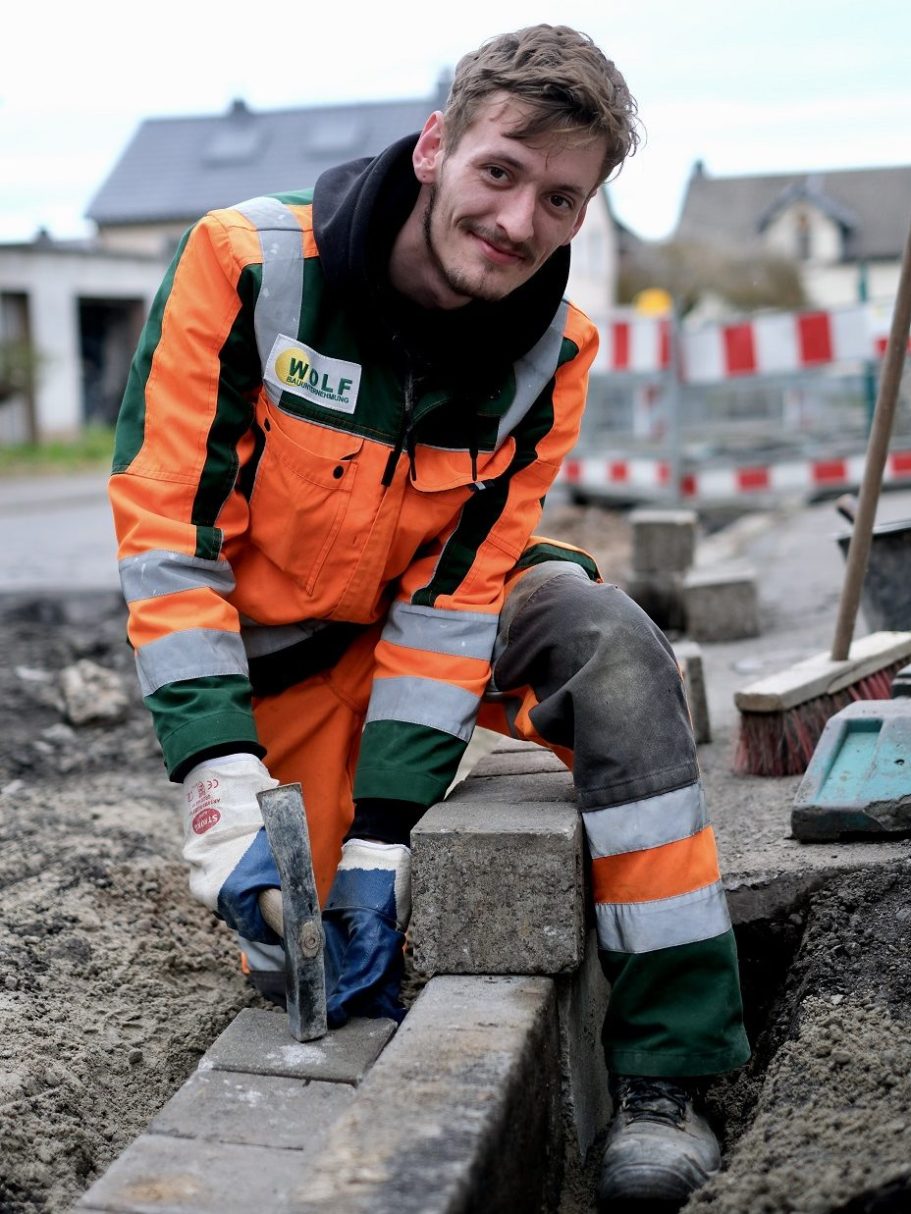 Straßenbau im Tiefbau: Bordsteinabsenkung für Grundstückszufahrt Ein lächelnder Straßenbauer in Arbeitskleidung setzt mit dem Hammer Pflastersteine an einer Bordsteinabsenkung