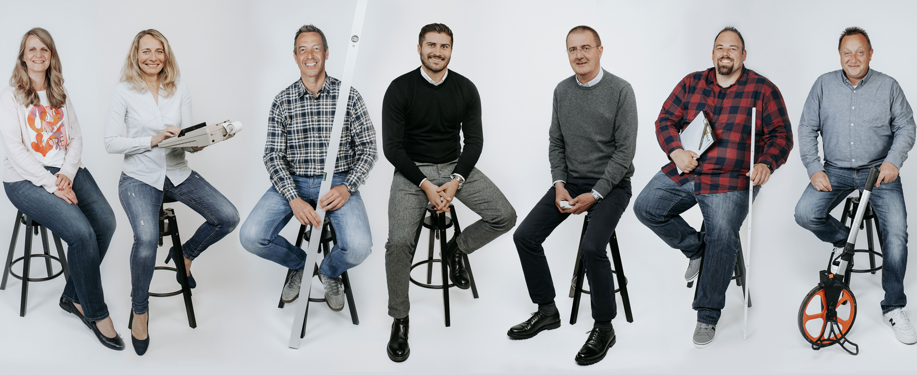 Sechs Personen sitzen auf Hockern, lächelnd, in lockerer Kleidung vor neutralem Hintergrund. Es ist das Büro Team der Firma