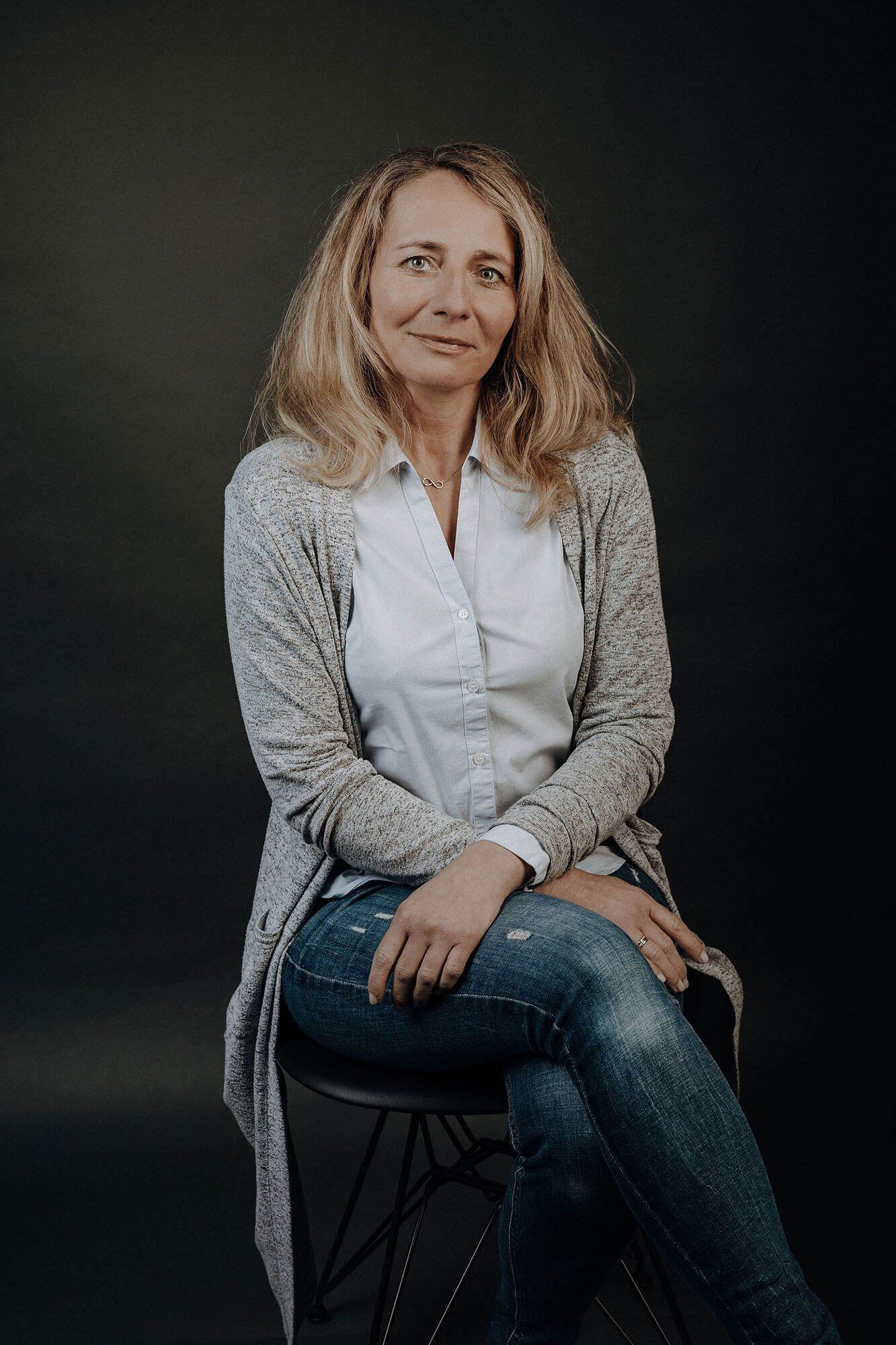 Frau mit blonden Haaren sitzt auf einem Stuhl, lächelt, trägt eine Bluse und Jeans.