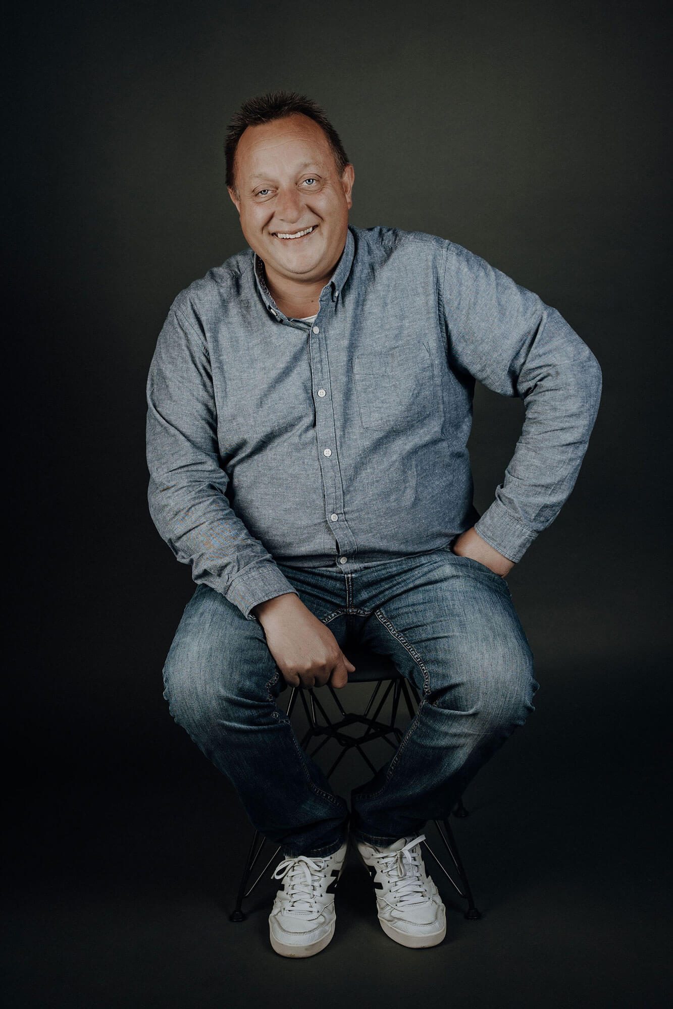 Lächelnder Mann in einem blauen Hemd und Jeans, mit einer Hand auf dem Knie.