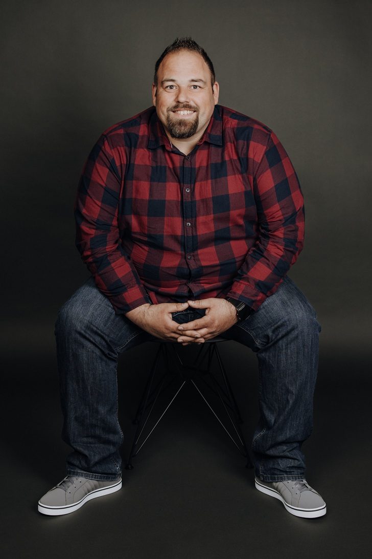 Lächelnder Mann in einem blauen Hemd und Jeans, mit einer Hand auf dem Knie.