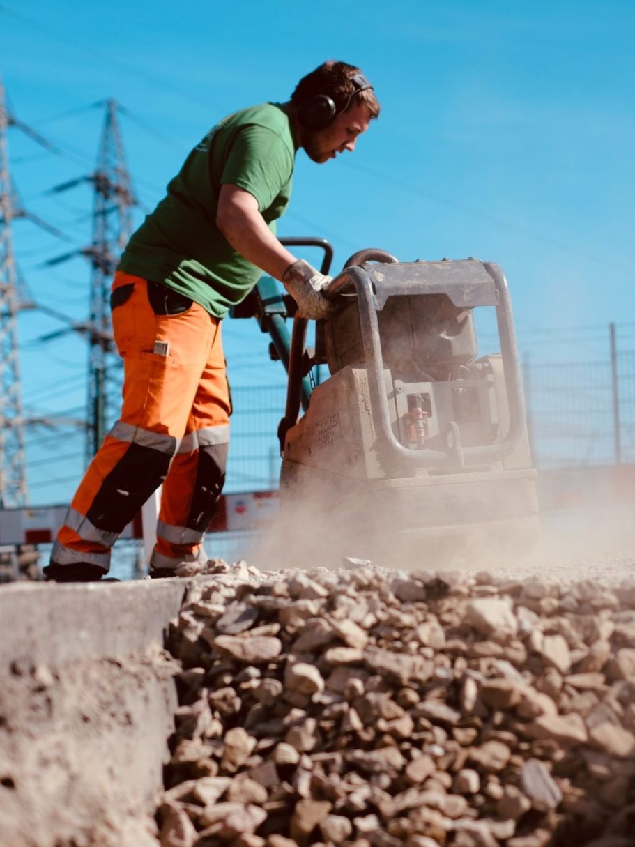 Tiefbauarbeiten: Bodenverdichtung mit der Rüttelplatte Bauarbeiter der Karl Wolf GmbH & Co. KG in Arbeitskleidung verdichtet mit einer Rüttelplatte den Schotterunterbau auf einer Tiefbaubaustelle