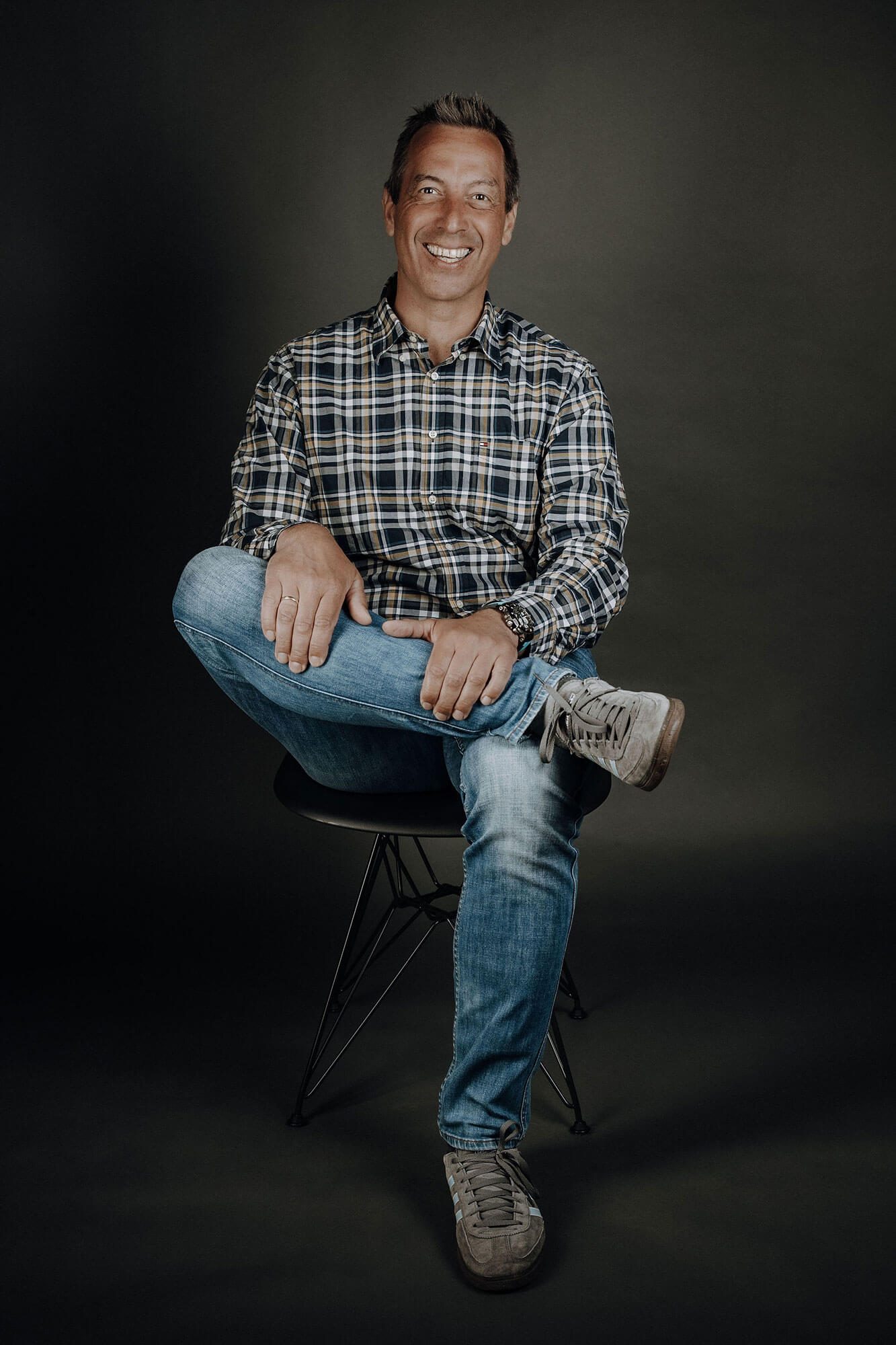 Mann sitzt lächelnd auf einem Stuhl, trägt ein kariertes Hemd und Jeans.