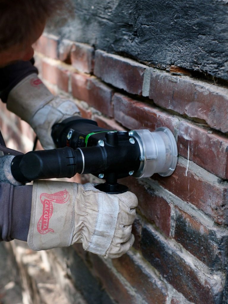 Fachgerechtes Ausfräsen von Mauerwerksfugen zur Sanierung Nahaufnahme eines Handwerkers mit Handschuhen, der eine Elektrofräse mit Staubabsaugung verwendet, um alte Fugen aus einer Ziegelsteinmauer