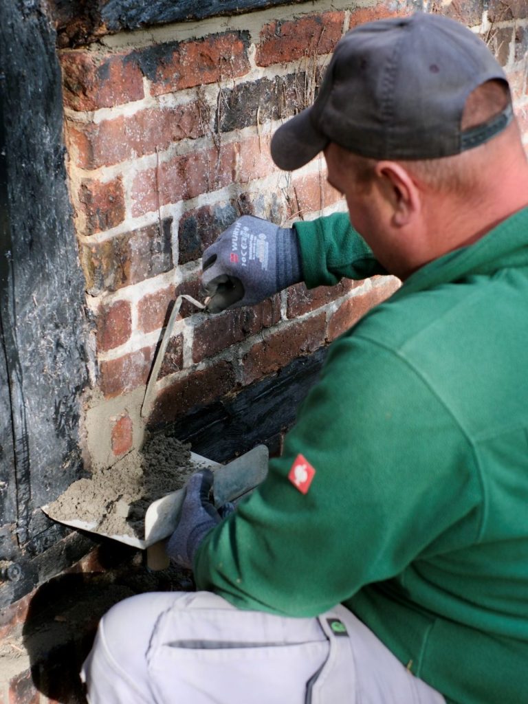 Fachgerechtes Einbringen des Spezialmörtels mit dem Fugeneisen Handwerker bringt mit Fugeneisen und Mörtelbrett frischen Sanierungsmörtel präzise in die vorbereiteten Fugen einer Klinkerwand ein.